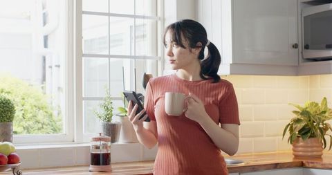 Asian woman in kitchen holding smartphone and mug in sunny kitchen