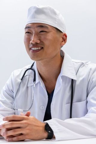 Asian doctor smiling while seated at clinic table wearing lab coat, cap and stethoscope