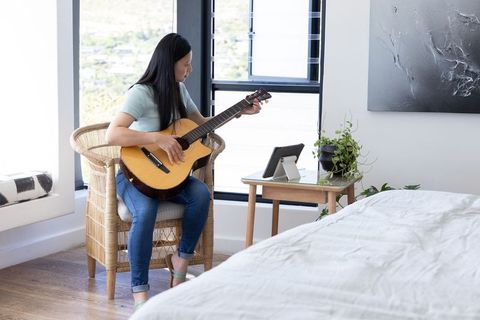 Mature woman practicing acoustic guitar in bright minimalist bedroom with tablet lesson