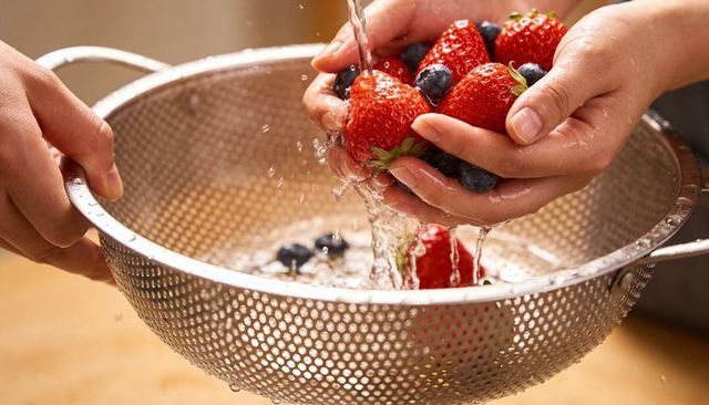 Hands rinsing fresh strawberries and blueberries over stainless colander, water droplets shining