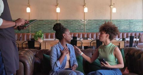 African american friends chatting on leather sofa in cozy modern cafe holding smartphone