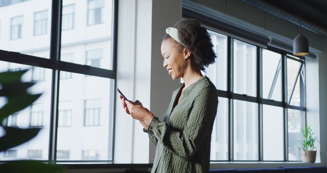 Woman is intently working on her smartphone while standing in a modern office filled with natural daylight. Useful for illustrating themes like professional communication, workplace technology adoption, and focused productivity. Ideal for business, corporate training, or tech industry content.
