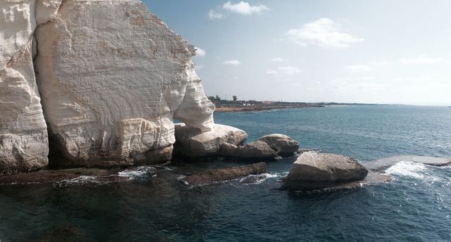 Massive white limestone cliff against a beautiful shoreline with rocky outcrops. The clear sky and sunny weather highlight the calm blue ocean waters. Ideal for promoting travel destinations, beach tourism, outdoor activities, nature preservation, and landscape photography.
