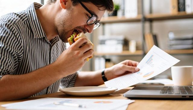 Young man eating sandwich while reviewing business charts at desk with laptop