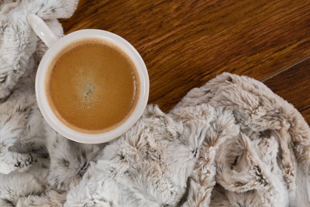 Close-up of a coffee cup placed on a wooden table next to a soft, fluffy blanket. Ideal for themes related to comfort, relaxation, home interiors, and cozy mornings. Perfect for use in blogs, social media posts, and advertisements promoting warm beverages, home decor, and lifestyle content.