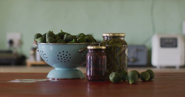 Image shows fresh figs in a colander alongside jars of pickled figs on a kitchen countertop. Perfect for use in culinary blogs, recipes, healthy eating articles, and promotional materials for food preservation-related products.