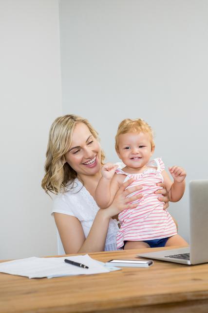 Mother holding baby girl on her lap while using a laptop at home. Both are smiling and appear to be in a home office setting. Ideal for illustrating concepts of work-life balance, parenting, remote work, and family bonding. Suitable for articles, blogs, and marketing materials about work-from-home parents, multitasking, and childcare solutions.
