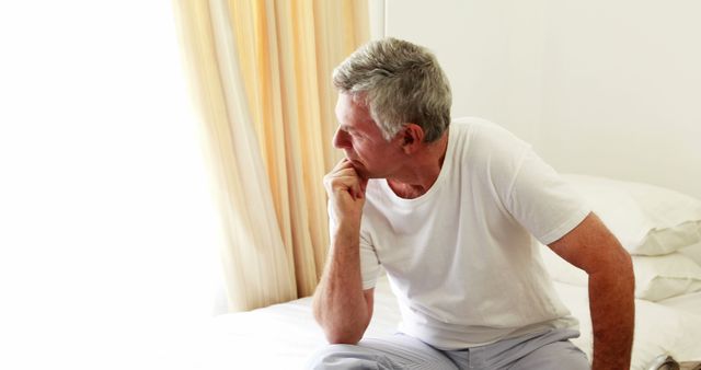 Elderly man with gray hair sitting on bed, staring thoughtfully out of bright window. Ideal for themes related to aging, contemplation, relaxation, and home life. Useful in articles or advertisements focusing on senior mental health, lifestyle, wellbeing, or personal reflection.