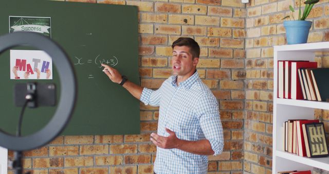 Male math teacher providing online teaching to students via livestream. He is explaining algebraic equations on a chalkboard, geared up for a virtual classroom setting. Ideal for illustrating remote education, e-learning concepts, online tutorials, and modern teaching methods.