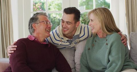 Adult son embracing smiling parents on cozy living room sofa, enjoying family moment