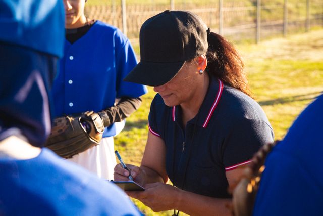 Female baseball coach taking notes while discussing game tactics with her team. Use this for sports training articles, coaching strategies, team building exercises, or promotional material for women's sports leagues.