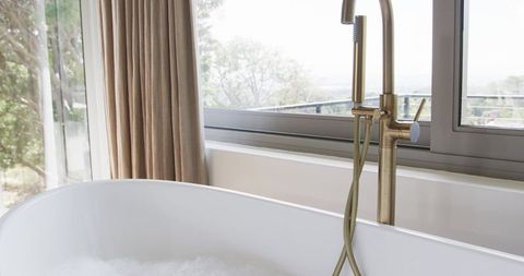 Freestanding white bathtub with brass faucet bubbling with foam by window luxury spa style