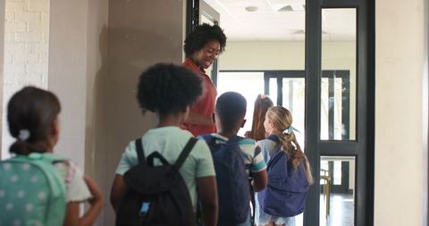 Teacher greeting students at school entrance carrying backpacks and smiling