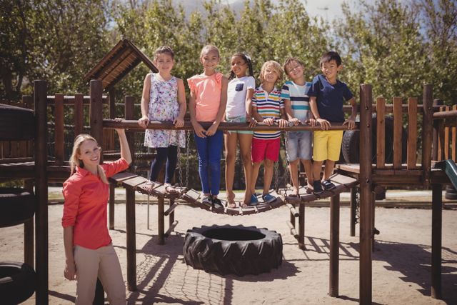 Group of cheerful schoolchildren standing on a playground bridge with their teacher. The children are smiling and appear to be enjoying their time outdoors. Ideal for use in educational materials, advertisements for schools or playground equipment, and articles about childhood development and outdoor activities.