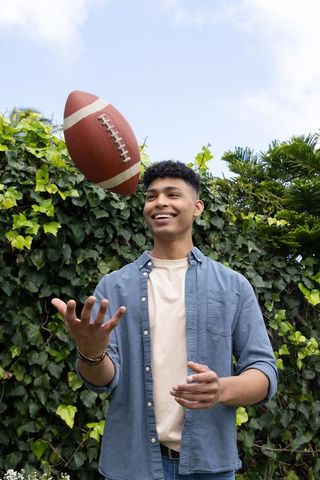 Young man tossing football in sunny backyard garden wearing denim shirt and smiling