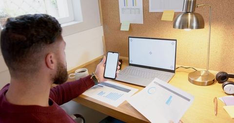 Bearded man analyzing charts at home desk using smartphone and laptop for remote work