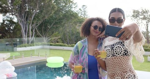 Female friends taking selfie poolside while holding drinks and wearing summer cover-ups