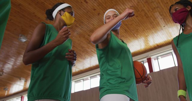 Female basketball players wearing masks are celebrating on a basketball court. They are demonstrating team spirit and unity in a sports hall with a wooden ceiling. This image is perfect for use in articles or advertisements focusing on sportsmanship, safety in sports, health protocols in athletics, teamwork, or youth sports activities.