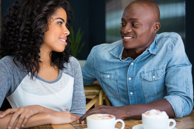 Young couple sitting at a table in a coffee shop, smiling and looking at each other. They are enjoying their time together, indicating a romantic relationship. This image can be used for advertisements, blog posts, or articles about dating, relationships, or social activities.