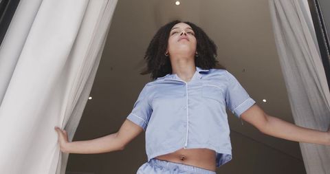 Standing in doorway wearing light blue pajamas opening curtains to morning light