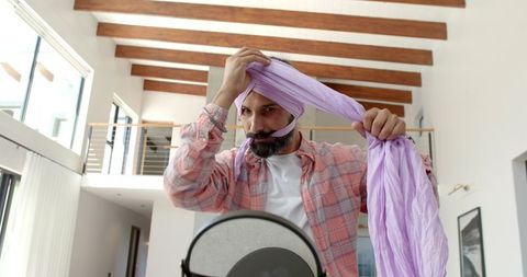 Mid-adult indian man tying lavender turban in modern loft using vanity mirror