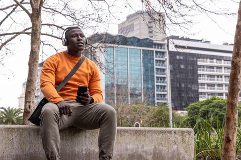African american man listening to music on headphones holding smartphone in city plaza