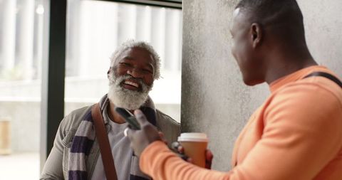 Two african american men laughing and chatting in transit lobby holding coffee and phone