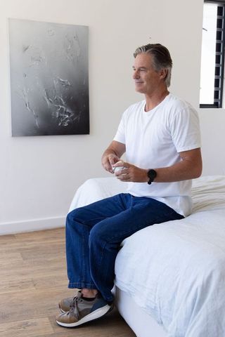Mature man sitting on bed edge holding round glass in bright minimalist bedroom