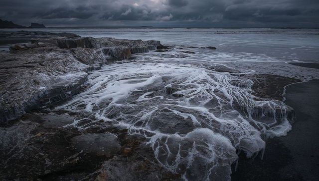 Lace-like sea foam threading over rocky shoreline at low tide under stormy sky
