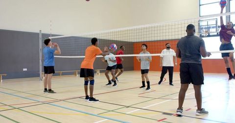 Multiethnic male team playing indoor volleyball jumping to spike and block on gym court