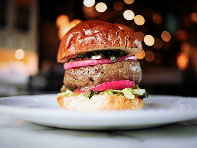 Gourmet beef burger with bun, onions, herbs, and lettuce served on white plate in restaurant. Perfect for use in menu designs, food blogs, restaurant promotions, and culinary magazines.