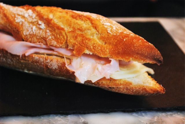 A ham and cheese baguette displayed on a black surface, showcasing the fresh ingredients and classic combination of flavors. Perfect for illustrating concepts related to savory meals, lunches, snacks, deli products, or artisanal sandwiches. Great for use in food blogs, culinary websites, menu designs, and social media posts promoting bakery or deli shop offerings.