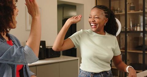 Laughing diverse female friends high-fiving in kitchen celebrating joyful lifestyle moment