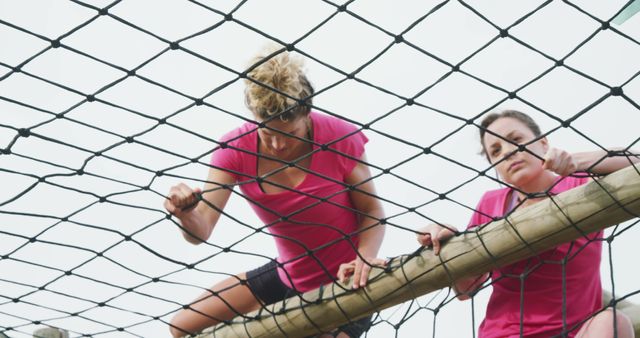 Two women climbing rope net, focused on overcoming an obstacle during a race. Ideal for use in content promoting teamwork, fitness motivation, outdoor activities, sports events, and determination.