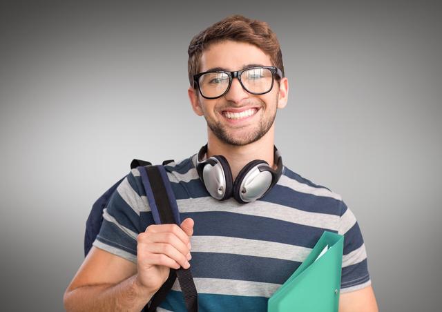 This stock photo captures a young student brightening a gray background with his smile. He looks ready for class as he holds a notebook and wears a backpack along with headphones and glasses. Perfect for educational websites, school promotional materials, tutoring services advertisements, and motivational academic content.