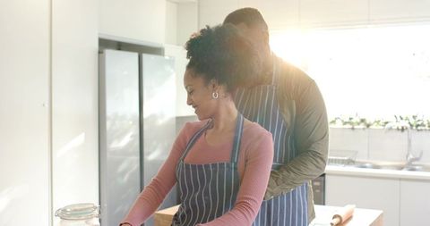 Sunlit modern kitchen couple baking together, man hugging woman at island in striped aprons
