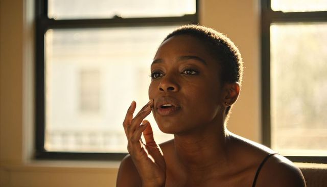 Low-light portrait of contemplative black woman touching face near sunlit window