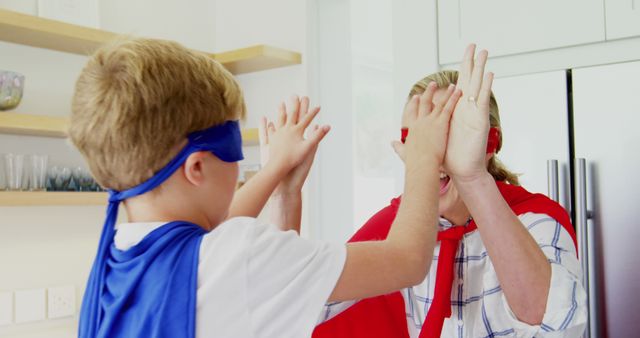 Two Caucasian children are playing in a kitchen, one wearing a blue superhero mask and cape, and the other with a red mask and cape, with copy space. They appear to be engaged in a playful pretend battle, adding a sense of imagination and fun to the domestic setting.