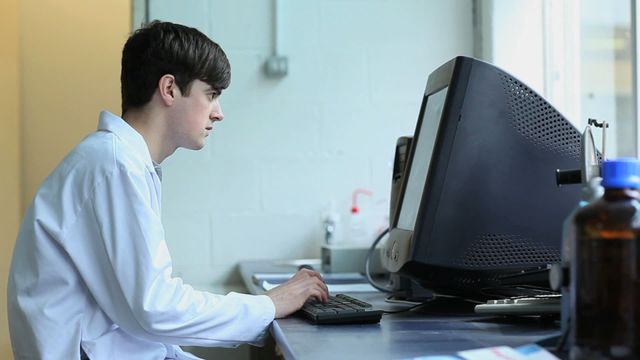 Young scientist wearing lab coat, focused on computer screen while working in a modern laboratory. Ideal for illustrating themes of scientific research, laboratory work, young professionals, technology in science, and modern lab settings. Can be used on websites, blogs, and articles related to science, research, and technology in laboratories.