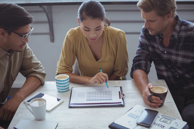 Three young professionals are gathered around a table in an office, discussing and planning a project. They are looking at documents and charts while enjoying coffee. This image can be used for business and teamwork concepts, professional development, and collaborative work environments.