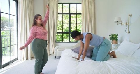 Multiracial women dancing and smoothing pillow in bright cozy bedroom morning lifestyle