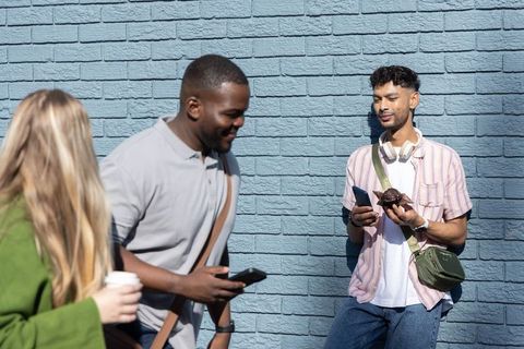 Sunlit diverse young adults walking past blue brick wall, man leaning holding phone and pastry
