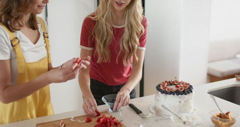 Friends decorating layered cake with fresh figs and strawberries in bright home kitchen