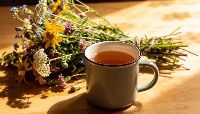 Sunlit enamel mug of herbal tea with wildflower bouquet on wooden kitchen table, cozy
