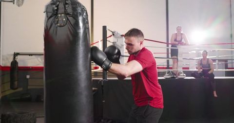 Male boxer training on heavy bag with women watching from ring, backlit gym scene, fitness