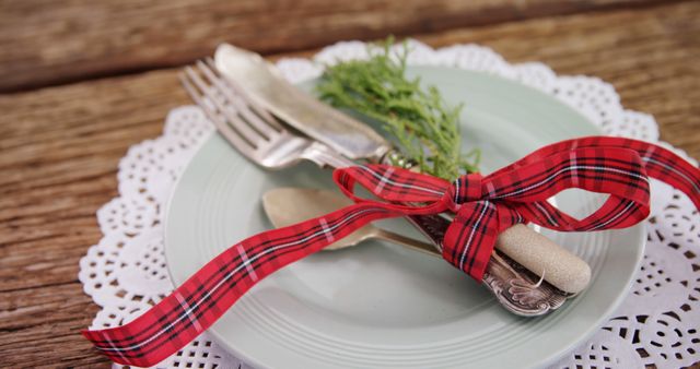 A set of silverware is tied with a festive red plaid ribbon on a pastel green plate, with a sprig of greenery adding a touch of elegance. The arrangement suggests a holiday or celebratory meal, with attention to detail in the presentation.