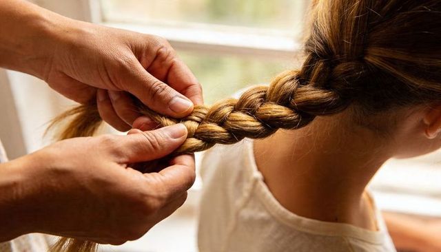 Caregiver braiding child hair at window — intimate family morning routine