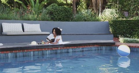 Relaxing poolside reading woman lounging on wooden deck with book, hat and beach ball