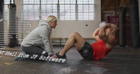 Kneeling coach guiding athlete doing crunches on gym floor with boxing bags and windows