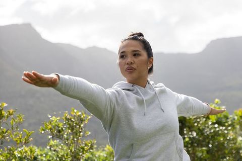 Mid-adult asian woman stretching arms in mountain valley wearing light gray hoodie